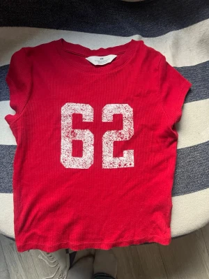 Röd t-shirt med siffran 62 från H&M - Cool röd t-shirt från H&M med trycket '62' framtill i vitt. T-shirten har korta ärmar och är gjord i ribbad bomull som ger en skön och avslappnad känsla. Perfekt för dig som gillar sportig stil och vill sticka ut lite extra.