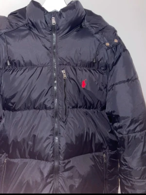 Svart polo  pufferjacka  - Snygg svart pufferjacka med huva och dragkedja framtill. Jackan har en röd broderad logga på bröstet, flera praktiska fickor med dragkedja och justerbar huva. Materialet är glansigt och quiltat för extra värme och stil.