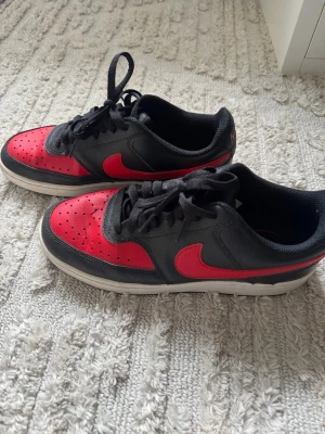 Svarta och röda Nike sneakers - Snygga sneakers från Nike i svart och rött med klassisk låg siluett. Skorna har svart ovandel i läder med röda detaljer och swoosh, samt vit sula. Perfekta för dig som gillar streetwear och vill sticka ut med färg.