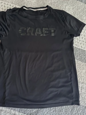 Svart t-shirt från Craft JR  152-158 cm  - Snygg svart t-shirt från Craft med stor logga framtill. Tillverkad i ett lätt och funktionellt syntetmaterial som passar perfekt för träning. Klassisk rund halsringning och korta ärmar för en sportig look.