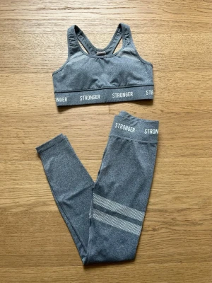 Grått träningsset från Stronger - Snyggt grått träningsset från Stronger med sport-bh och matchande tights. Setet har vita logotyper och diskreta mönsterdetaljer på benen. Materialet är stretchigt och passar perfekt för träning eller chill. Sport-bhn har brottarrygg för extra stöd.
