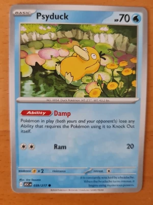 Pokémonkort Psyduck 039/217 - Säljer ett Pokémonkort med Psyduck från Sword & Shield-serien, nummer 039/217. Kortet har blå ram, färgglad illustration av Psyduck vid en damm och detaljer om attackerna Damp och Ram. Perfekt för samlare eller Pokémonfans!