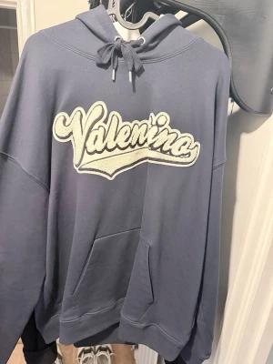 Blå hoodie med Valentino-tryck - Säljer en blå hoodie med stor vit Valentino- text på bröstet. Perfekt för dig som gillar streetwear och vill ha en chill look.
