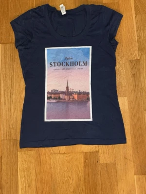 Mörkblå t-shirt med Stockholm-tryck - Snygg mörkblå t-shirt med ett stort tryck av Stockholm på framsidan. T-shirten har rund halsringning och korta ärmar. Materialet känns mjukt och skönt, troligtvis bomull eller en bomullsblandning. Perfekt för dig som gillar city vibes och vill ha något unikt i garderoben.