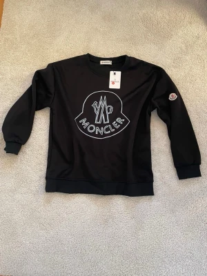 Svart sweatshirt från Moncler - Svart sweatshirt från Moncler med stort vitt logotryck på bröstet och Moncler-märke på ärmen. Tröjan har rund halsringning, ribbade muddar och är gjord i mjukt material som känns skönt mot huden. Perfekt för en avslappnad och trendig look.