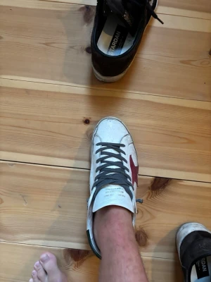 Vita sneakers med röd stjärna - Säljer ett par vita sneakers med grå snörning och en röd stjärna på sidan. Skorna har även gröna detaljer vid hälen och svart text på sidan. Klassisk låg modell som passar till det mesta.