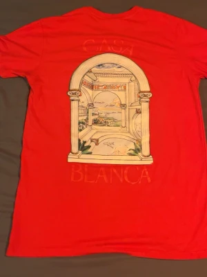 Röd Casablanca t-shirt med tryck - Säljer en röd t-shirt från Casablanca med ett stort grafiskt tryck av en klassisk arkad och landskap på ryggen samt ett mindre tryck på bröstet. T-shirten har rund hals och korta ärmar. Perfekt för dig som gillar unika prints och färg.