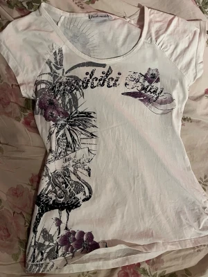 Vit t-shirt med blommigt tryck Fresh Made - Säljer en vit t-shirt från Fresh Made med blommigt tryck i svart och lila samt texten 'Waikiki Bay' framtill. T-shirten har korta ärmar och rund halsringning. Perfekt för dig som gillar unika detaljer och sköna material.