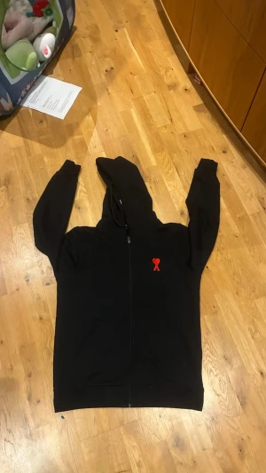 Ami tröja - Säljer en svart hoodie med dragkedja och huva. På bröstet finns ett broderat rött hjärta med band, vilket ger en unik detalj. Hoodien har klassisk passform och är perfekt för en chill och avslappnad stil. Materialet känns mjukt och bekvämt. perfekt till dig som är i storlek S/M 175cm+ Pris kan diskuteras