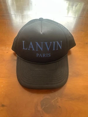 Svart lanvin keps  - Snygg svart truckerkeps från Lanvin Paris med blå text framtill. Kepsen har mesh på sidorna och justerbar snapback baktill för perfekt passform. Perfekt accessoar för en trendig och avslappnad stil.