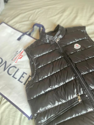 Svart dunväst från Moncler - Snygg svart dunväst från Moncler med glansig finish och klassisk logga på bröstet. Västen har hög krage, två dragkedjeförsedda fickor och en stilren, quiltad design. Perfekt för lager-på-lager och streetwear-vibe.