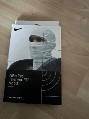 Nike Pro Therma-FIT Hood balaclava - Nike Pro Therma-FIT Hood är en vit balaclava med svart Nike-logga. Tillverkad i ett värmande material som hjälper till att hålla värmen. Snygg, tight passform som kan bäras under hjälm. Perfekt för kalla dagar och sportaktiviteter.