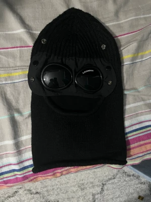 Svart balaclava med goggles C.P. Company - Säljer en svart balaclava från C.P. Company med inbyggda goggles och öppning för munnen. Tillverkad i stickat material och har detaljer i plast runt glasögonen. Perfekt för dig som vill sticka ut med en unik accessoar.