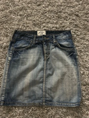 Blå jeanskjol med fickor - Säljer en klassisk blå jeanskjol i kort modell med slitna detaljer och fem fickor. Kjolen har knapp och dragkedja framtill samt två bakfickor med knapp. Perfekt till vardags för en avslappnad look.