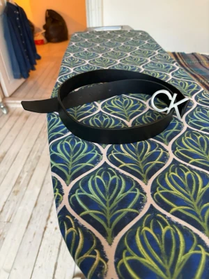 Svart bälte med CK-spänne - Snyggt svart bälte från Calvin Klein med ett stilrent metallspänne i form av CK-loggan. Bältet är i slätt skinn och passar perfekt till jeans eller byxor för en clean och modern look.