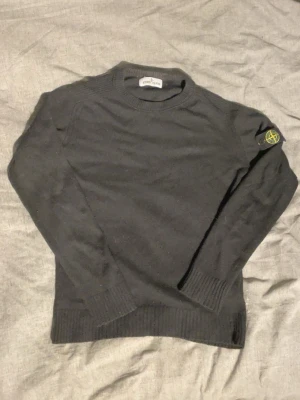 Svart stickad tröja från Stone Island - Snygg svart stickad tröja från Stone Island med den klassiska loggan på vänster ärm. Tröjan har rund halsringning, ribbade muddar och är tillverkad i ett mjukt material som passar perfekt till höst och vinter. Pris kan diskuteras 