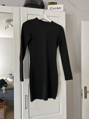 Svart långärmad kort klänning - En stilren svart klänning med lång ärm och rund hals. Klänningen är kort och har en figurnära passform som framhäver siluetten. Perfekt för dig som gillar minimalistisk och enkel stil.