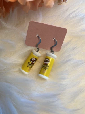 Pringles örhängen i gult - Unika örhängen formade som små Pringles-rör i gul färg med detaljerad logga och text. Hängena är tillverkade i plast och har metallkrokar. Perfekt för dig som gillar roliga och annorlunda accessoarer.