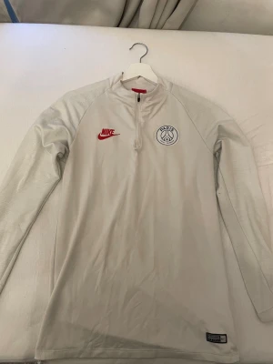 Vit Nike PSG träningströja - Snygg vit långärmad träningströja från Nike med PSG-logga på bröstet och röd Nike-logga. Tröjan har halv dragkedja och är tillverkad i Dri-FIT-material som andas, perfekt för fotbollsträning eller sportaktiviteter.