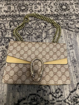  beige monogram handväska - Snygg beige handväska från  med klassiskt monogram, guldfärgad kedjerem och dekorativt spänne framtill. Väskan har en rektangulär form och insidan är ljusbeige. Perfekt accessoar för dig som gillar lyxig och tidlös design.
