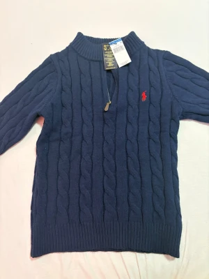 Polo Ralph lauren  - Mörkblå kabelstickad tröja från Polo Ralph Lauren med halv dragkedja och röd broderad logga på bröstet. Tröjan har ribbade muddar och klassisk passform. Perfekt för lager på lager och snygg till jeans.