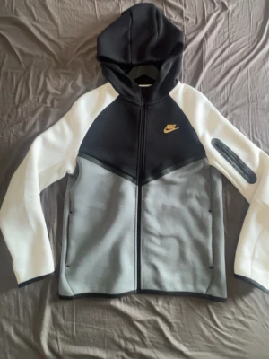 Nike tech fleece junior ,  i svart, grå och vit - Nike hoodie med dragkedja i svart, grå och vit färgblock. Gult Nike-märke på bröstet och praktisk huva. Två fickor framtill och en extra ficka med dragkedja på ena ärmen. Perfekt för en sportig och avslappnad stil.