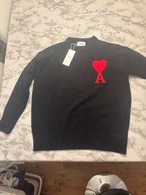 Svart stickad tröja från Ami - Snygg svart stickad tröja från Ami Alexandre Mattiussi med röd hjärtlogga och bokstaven A på bröstet. Tröjan har rund halsringning och ribbade muddar vid ärmslut och nederkant. Perfekt för dig som gillar stilrena detaljer och trendig design.