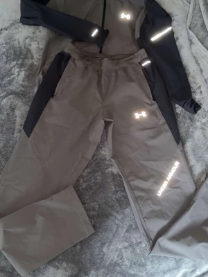 Under Armour träningsset M - Säljer ett sportigt träningsset från Under Armour i grått och svart. Setet består av jacka och byxor med reflekterande detaljer och dragkedjor vid fickorna. Perfekt för löpning eller utomhusträning. Materialet är lätt och snabbtorkande.