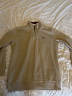 Boss tröja - Snygg beige sweatshirt från BOSS med halv dragkedja och ståkrage. Tröjan har diskret BOSS-logga på bröstet och cool detalj med randig insida på kragen i vitt och brunt. Perfekt för en clean och stilren look. Storlek S passar även som M