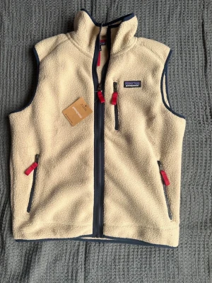 Beige fleeceväst från Patagonia - beige fleeceväst från Patagonia med marinblå detaljer och röda dragkedjor. Västen har hög krage, tre fickor med dragkedja och mjukt teddyfoder. Perfekt för lager-på-lager och outdoor vibes.