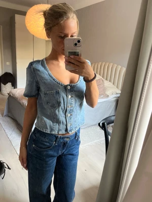 Ljusblå croppad jeanstopp - Säljer en ljusblå croppad jeanstopp med korta ärmar och v-ringning. Toppen har knappar framtill och en bröstficka. Perfekt till ett par höga jeans för en avslappnad och trendig look. Oanvänd!