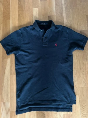 Svart pikétröja från Polo Ralph Lauren - Klassisk svart pikétröja från Polo Ralph Lauren med röd broderad logga på bröstet. Tröjan har korta ärmar, krage och knappar vid halsen. Tillverkad i mjuk bomull för en bekväm känsla och stilren look.