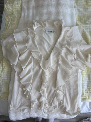 Krämvit volangblus från Kappahl - Supersöt krämvit blus från Kappahl med korta ärmar och volangdetaljer längs v-ringningen och framtill. Blusen är tillverkad i ett lätt, transparent tyg som ger en luftig känsla. Perfekt för en trendig och romantisk look.