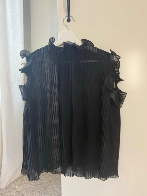 Plisserad svart blus med volanger - Snygg svart blus i plisserat, transparent tyg med volangdetaljer vid ärmöppningarna och halsen. Blusen är ärmlös och har en lös, flowy passform som ger en trendig och luftig look. Perfekt att styla med jeans eller kjol för en cool vibe.