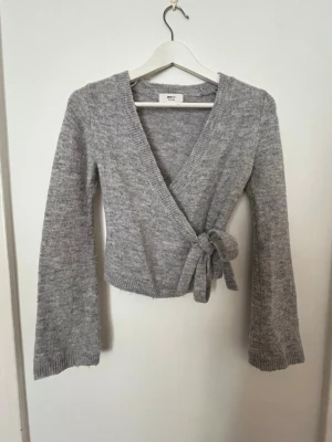 Grå omlottkofta från Gina Tricot - Mysig grå stickad omlottkofta från Gina Tricot i ullblandning. Koftan har långa ärmar, ribbade muddar och knyts i sidan för en snygg passform. 