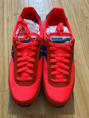 Saucony röda och blå sneakers - Snygga sneakers från Saucony i starkt röd färg med blå detaljer och vita inslag. Ovandelen är i textil och mocka, med klassisk Saucony-logga på sidan. Skorna har röda snören och en vit sula med svart yttersula för bra grepp.