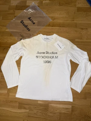 Vit långärmad t-shirt Acne Studios - Vit långärmad t-shirt från Acne Studios med tryck 'Acne Studios STOCKHOLM 1996' på bröstet. Klassisk passform och rund halsringning.           Storleken är M, men den är rätt så bägge av sig så L hade säkert också funkat. Den är helt oanvänd