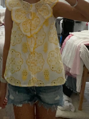 Gulvit ärmlös blus med broderi - Söt ärmlös blus i vitt med stora gula broderade blommönster och volangdetaljer vid axlarna. Blusen har en luftig passform och knäppning framtill. Perfekt för sommardagar och ger en fräsch och ungdomlig look.