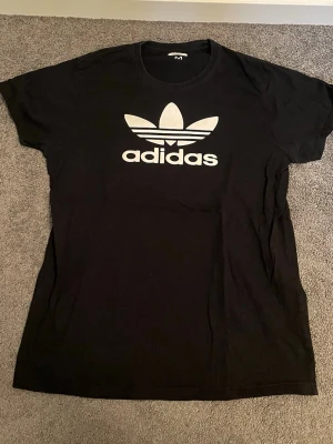 Svart Adidas t-shirt med logga - Säljer en svart t-shirt från Adidas med den klassiska vita trefoil-loggan och texten på bröstet. T-shirten har rund halsringning och korta ärmar. Perfekt för en sportig och avslappnad stil.