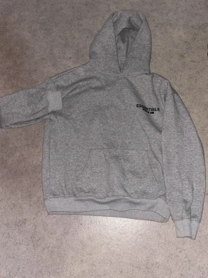 Grå Essentials Fear of God hoodie - Snygg grå hoodie från Essentials Fear of God med svart logga på bröstet och stor text på ryggen. Klassisk känguruficka framtill och huva. Perfekt för en avslappnad streetwear-look.