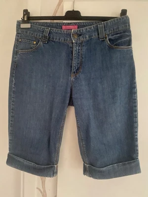 Blå jeansshorts med broderi - Säljer ett par klassiska blå jeansshorts med uppvikta benslut och snyggt mörkblått broderi på baksidan. Shortsen har fem fickor, bälteshällor och stängs med knapp och dragkedja. Perfekta för en avslappnad stil.