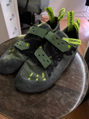 Gröna La Sportiva Tarantula klätterskor - Säljer ett par gröna La Sportiva Tarantula klätterskor med kardborreband och svarta detaljer. De har använts 5-6 gånger och är köpa i nya i butik. Skorna har slitstark gummisula och gula accenter på ovansidan. Perfekta för både inomhus- och utomhusklättring. 