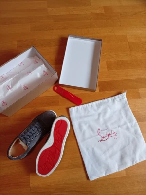 Gråa Louboutin sneakers med röd sula - Snygga gråa sneakers från Christian Louboutin med klassisk röd sula och vita kanter. Skorna har en låg profil, snörning framtill och är tillverkade i mocka. Perfekta för dig som vill sticka ut med en exklusiv touch.