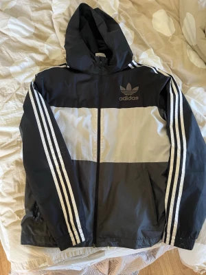 Svart och vit Adidas vindjacka - Snygg svart och vit vindjacka från Adidas med klassiska tre ränder längs ärmarna och huva. Jackan har dragkedja framtill, två sidofickor och Adidas-logga på bröstet. Perfekt för en sportig och avslappnad stil.