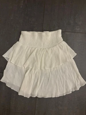 Vit volangkjol från SHEIN - Säljer en söt vit kortkjol från SHEIN med bred smock i midjan och dubbla volanger. Kjolen är gjord i ett lätt, skirt material som ger en flowy känsla. Perfekt för dig som gillar en feminin och trendig stil.