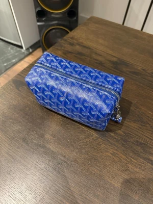 Blå mönstrad plånbok med dragkedja - Snygg blå plånbok med vitt grafiskt mönster och silverfärgad dragkedja. Rektangulär form med praktisk handledsrem. Perfekt för att hålla ordning på kort och kontanter. Materialet är syntet och ytan är lätt att torka av.