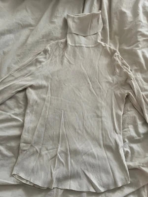 Beige ribbad polotröja från Zara - Säljer en beige ribbad polotröja från Zara. Tröjan har långa ärmar, smala ränder och en hög polokrage. Materialet är mjukt och stretchigt, perfekt för lager på lager. Enkel och stilren design som passar till det mesta.