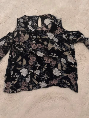Blommig svart blus med cut out-axlar - Snygg svart blus med blommigt mönster i vitt, beige och rosa toner. Blusen har cut out-axlar och lång ärm, samt en liten knapp i nacken. Tunt, luftigt material som ger en skön känsla. Perfekt för dig som gillar trendiga detaljer och blommigt.
