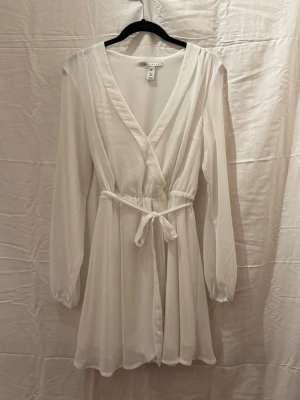 Vit omlottklänning med knytband - Snygg vit kort omlottklänning med v-ringning och knytband i midjan. Klänningen har långa ärmar och är tillverkad i ett lätt, luftigt material som ger ett elegant fall. Perfekt för dig som gillar en stilren och fräsch look.