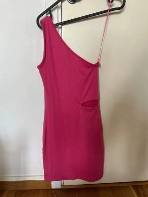 Rosa oneshoulder klänning Primark - Trendig rosa oneshoulder klänning från Primark i ribbad stretchig kvalitet. Klänningen har en smal axelrem och en cut-out detalj i sidan som ger en cool touch. Perfekt för dig som vill sticka ut med en färgstark look.